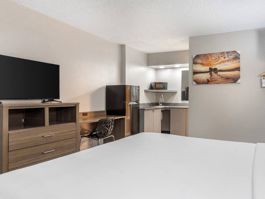 A hotel room with a bed, a TV, a kitchenette, and a painting on the wall.