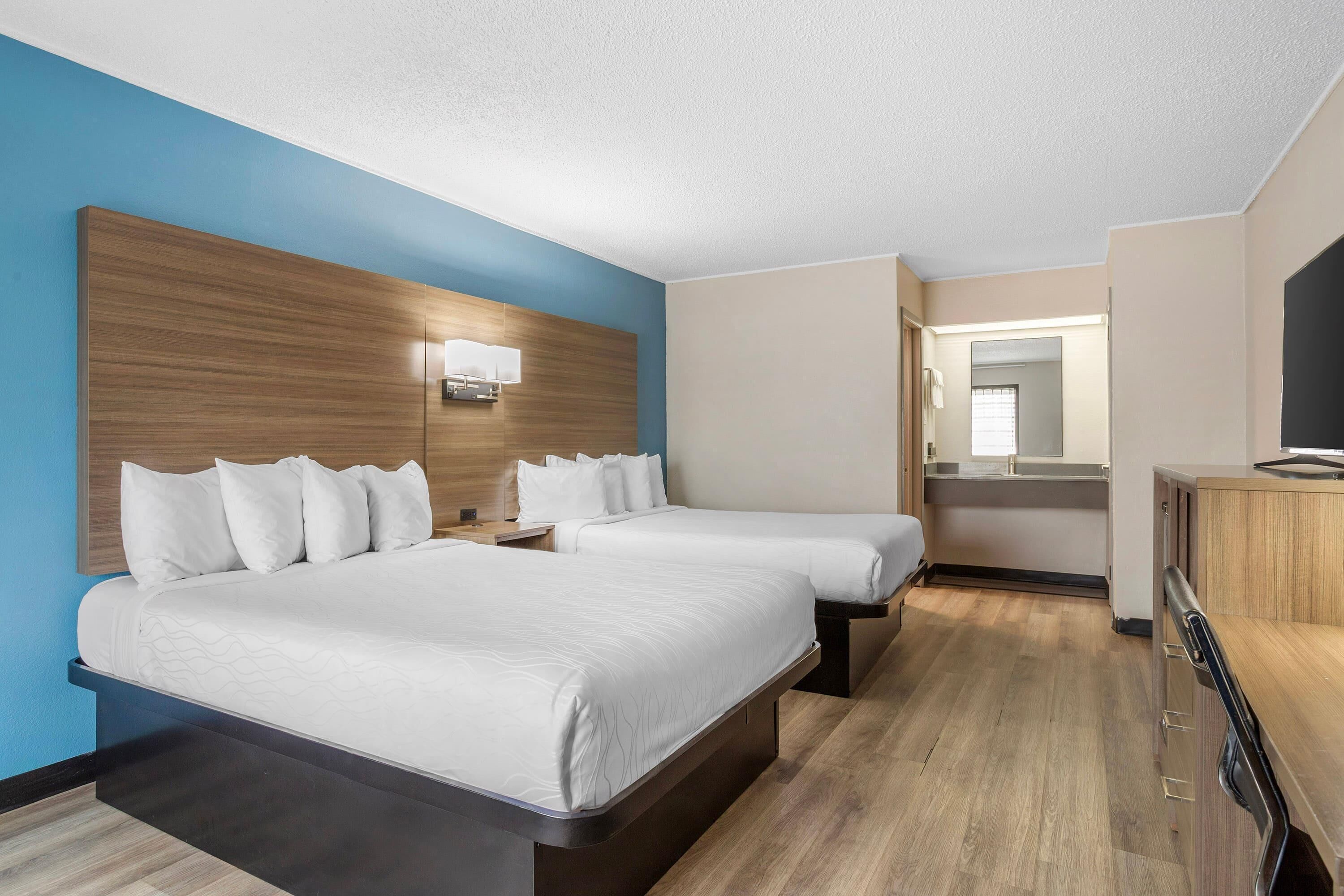 Ett hotellrum med två sängar, en sänggavel i trä, ett skrivbord med en stol, en TV och en spegel.