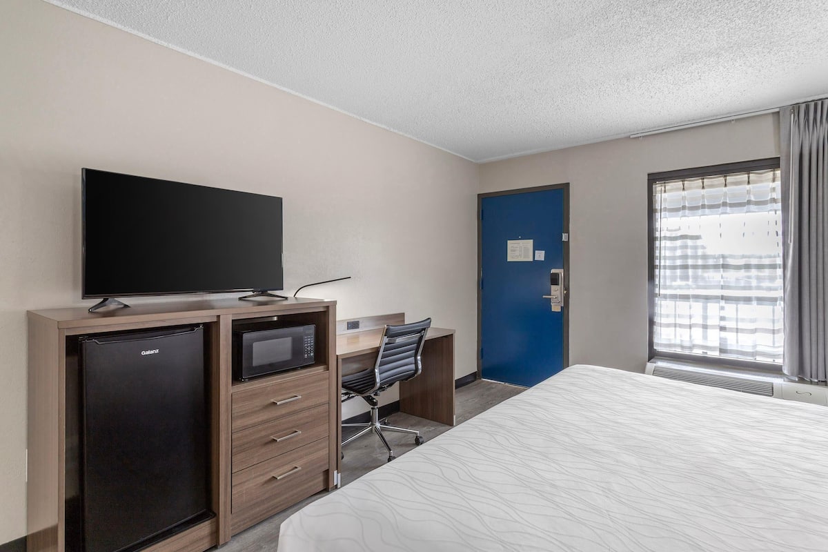 A hotel room with a bed, a desk with a chair, a television, a minibar, and a blue door.