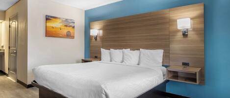 Une chambre d’hôtel moderne avec un grand lit, une tête de lit en bois, deux tables de chevet avec des lampes, un tableau au mur et une porte sur le côté gauche.