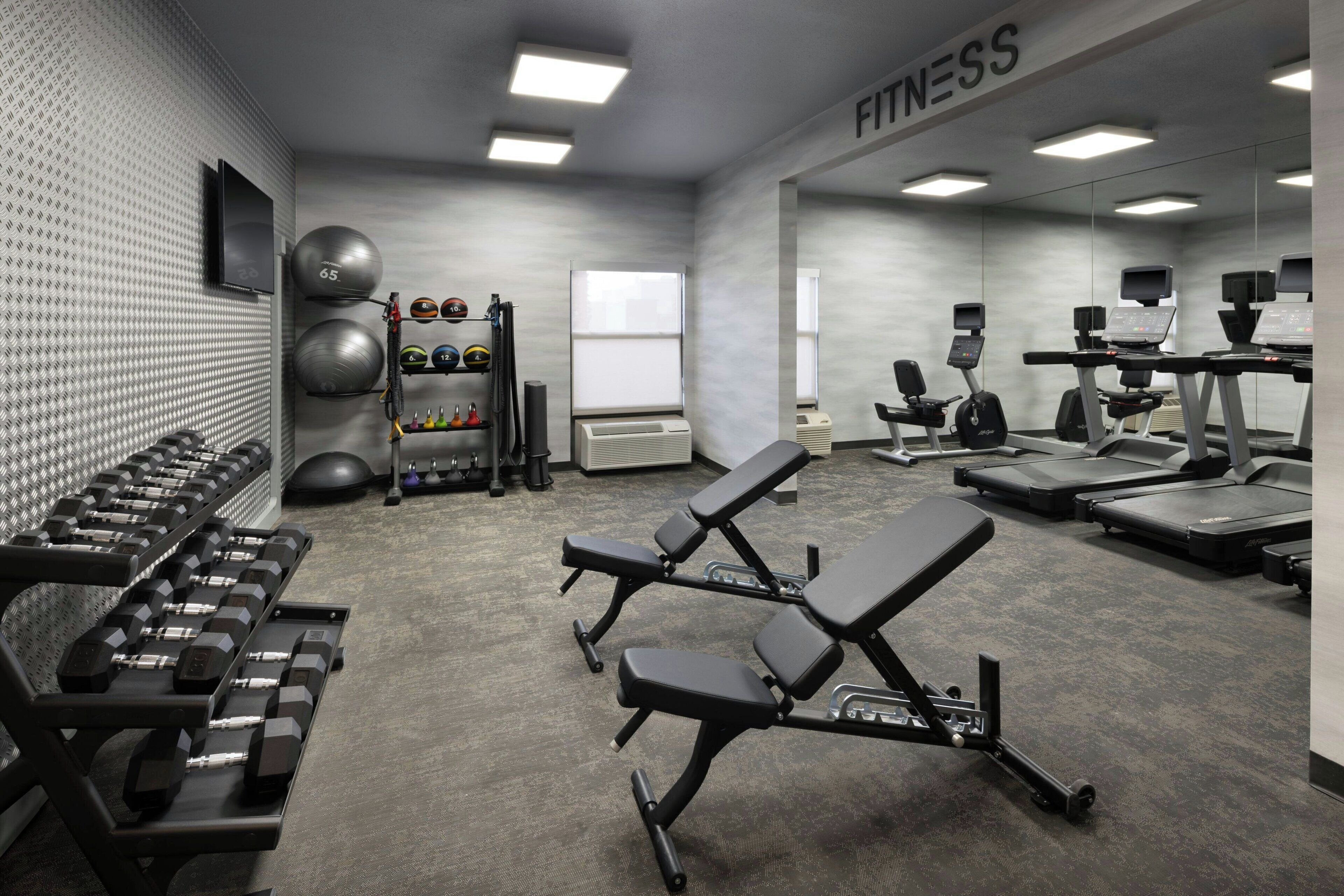 A gym with weight benches, dumbbells, exercise balls, treadmills, and a TV on the wall.