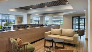 Un moderno vestíbulo de hotel con mostrador de recepción, zona de estar con sofá y sillones, y ventanas amplias con vistas al exterior.