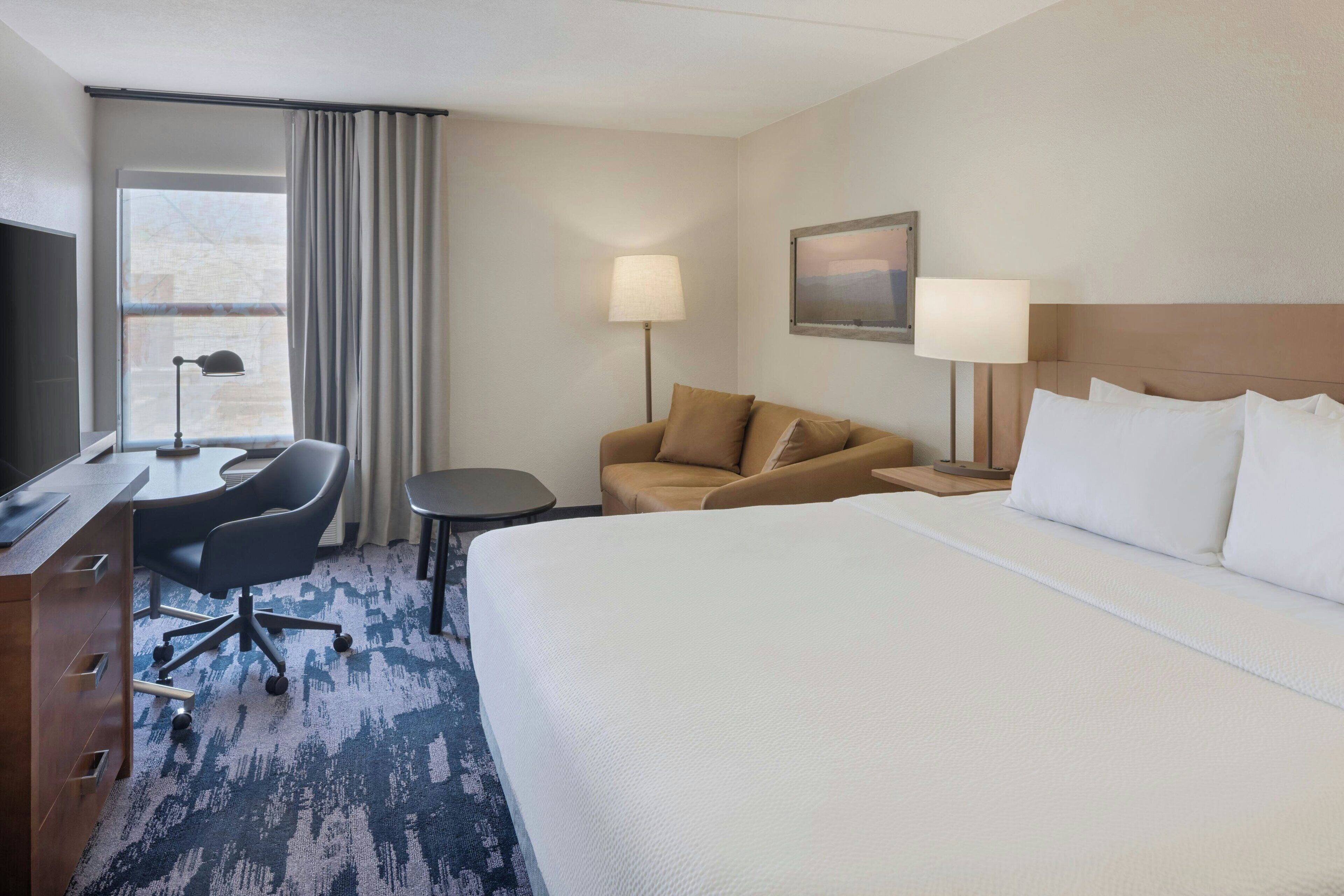 A hotel room with a large bed, a desk with a chair, a sofa, two bedside lamps, and a window with curtains.