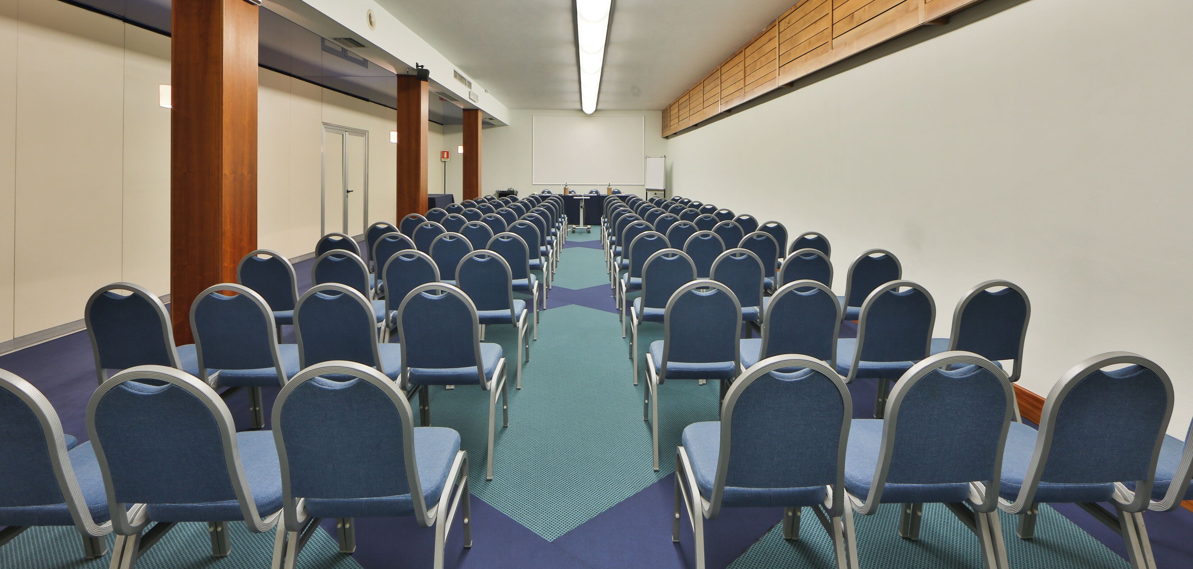 A conference room with rows of blue chairs, a projector screen, and a wooden panel on the wall.