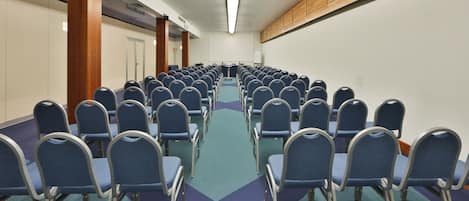 A conference room with rows of blue chairs, a projector screen, and a wooden panel on the wall.