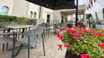 An outdoor seating area with tables and chairs, a potted plant with red flowers, and a building with arched windows in the background.