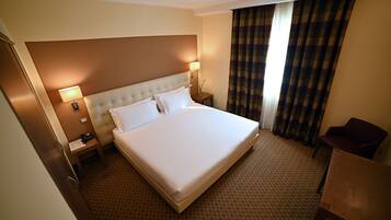 A hotel room with a large bed, two bedside lamps, a nightstand with a phone and papers, a chair, and a window with curtains.