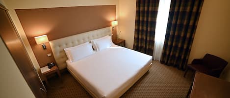 Habitación de hotel con una cama grande, dos lámparas de noche, una mesita de noche con un teléfono y documentos, una silla y una ventana con cortinas.