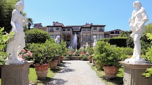A garden pathway flanked by statues leading to a building with a fountain.