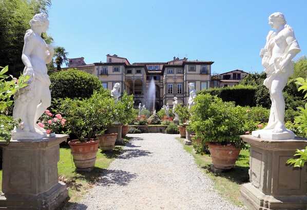 Un sendero en un jardín, bordeado por estatuas, que lleva a un edificio con una fuente.