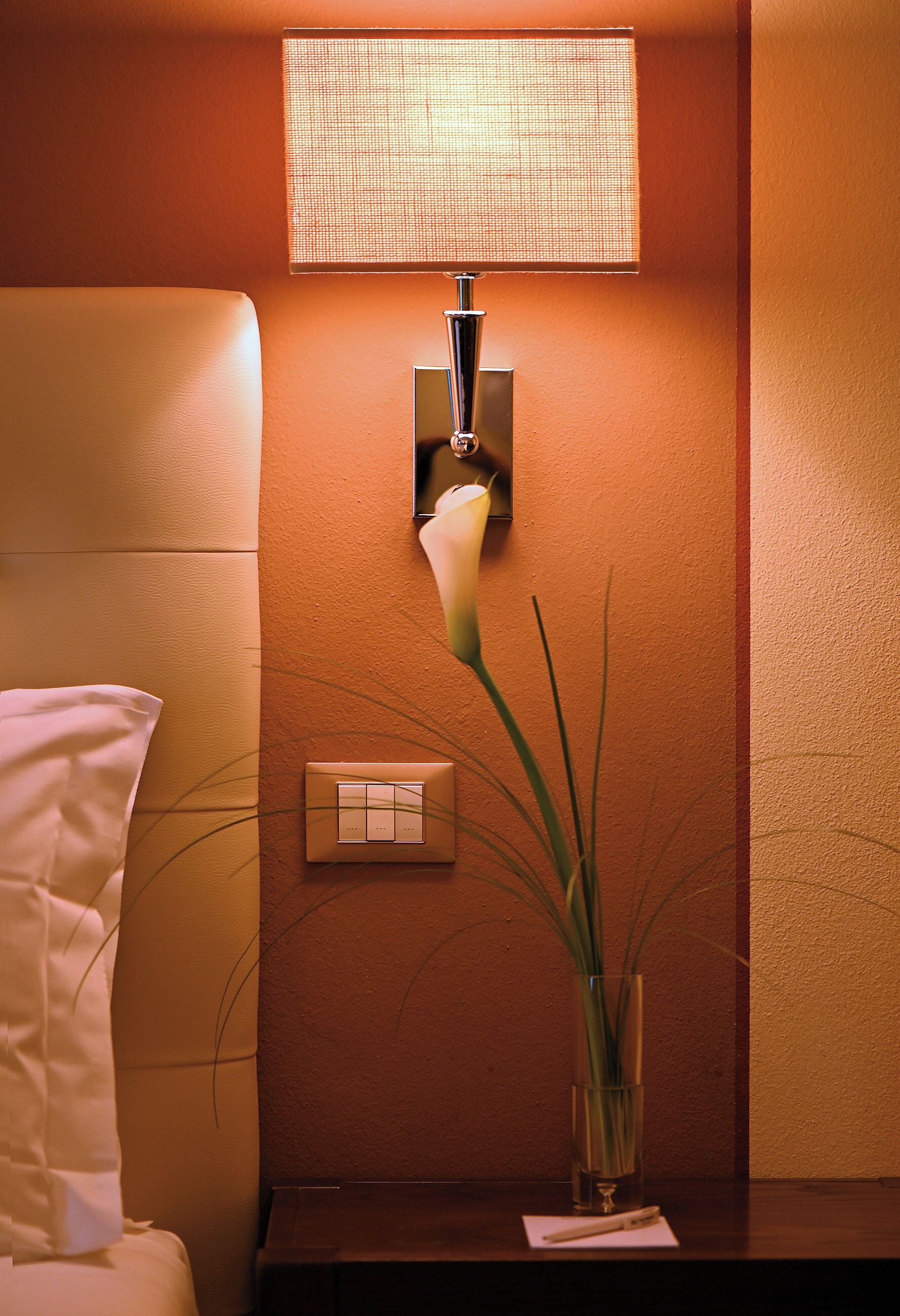 A hotel room with a bedside lamp, a bedside table with a vase of flowers, and a wall-mounted light switch.
