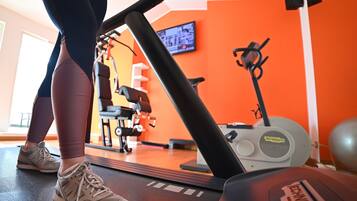 Ein Fitnessraum mit Trainingsgeräten, Laufband und Fernseher an der Wand.