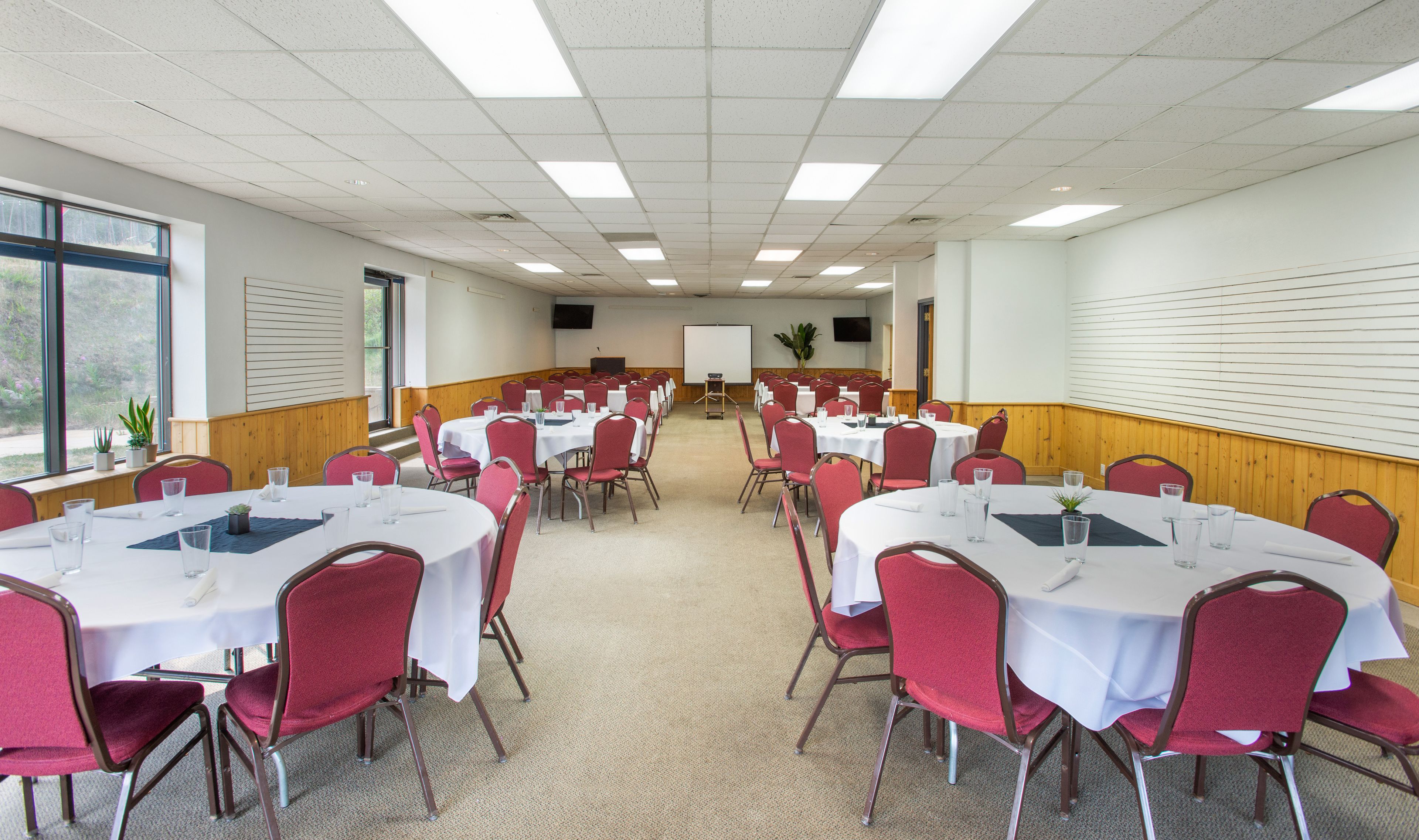 A conference room with multiple round tables and red chairs, equipped with white tablecloths and glass water glasses.