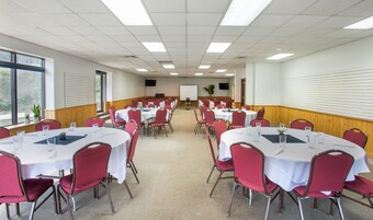 Un salón de conferencias con varias mesas redondas y sillas rojas, equipado con manteles blancos y vasos de vidrio para agua.