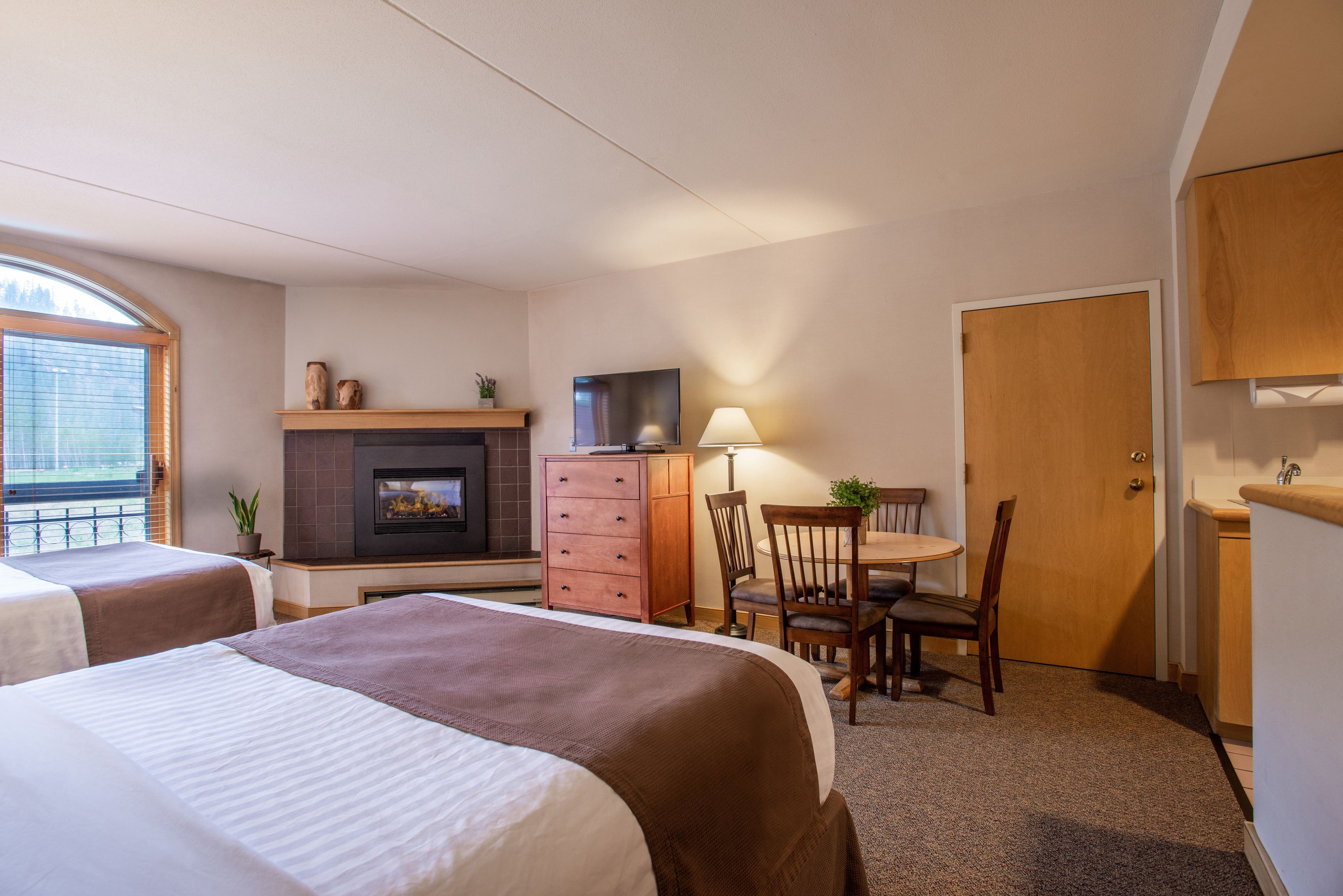 A hotel room with two beds, a fireplace, a television, a desk with a chair, and a door.
