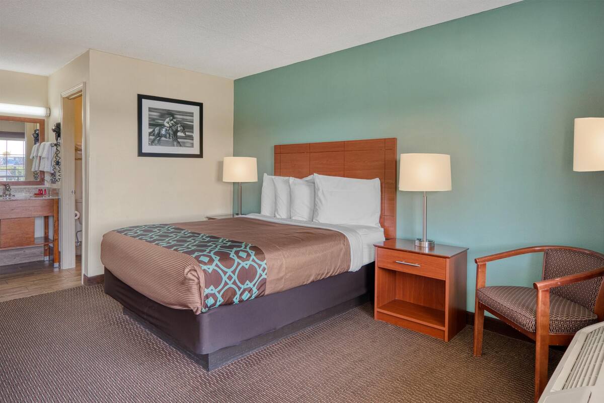 A hotel room with a bed, bedside lamps, a chair, a desk, and a framed picture on the wall.