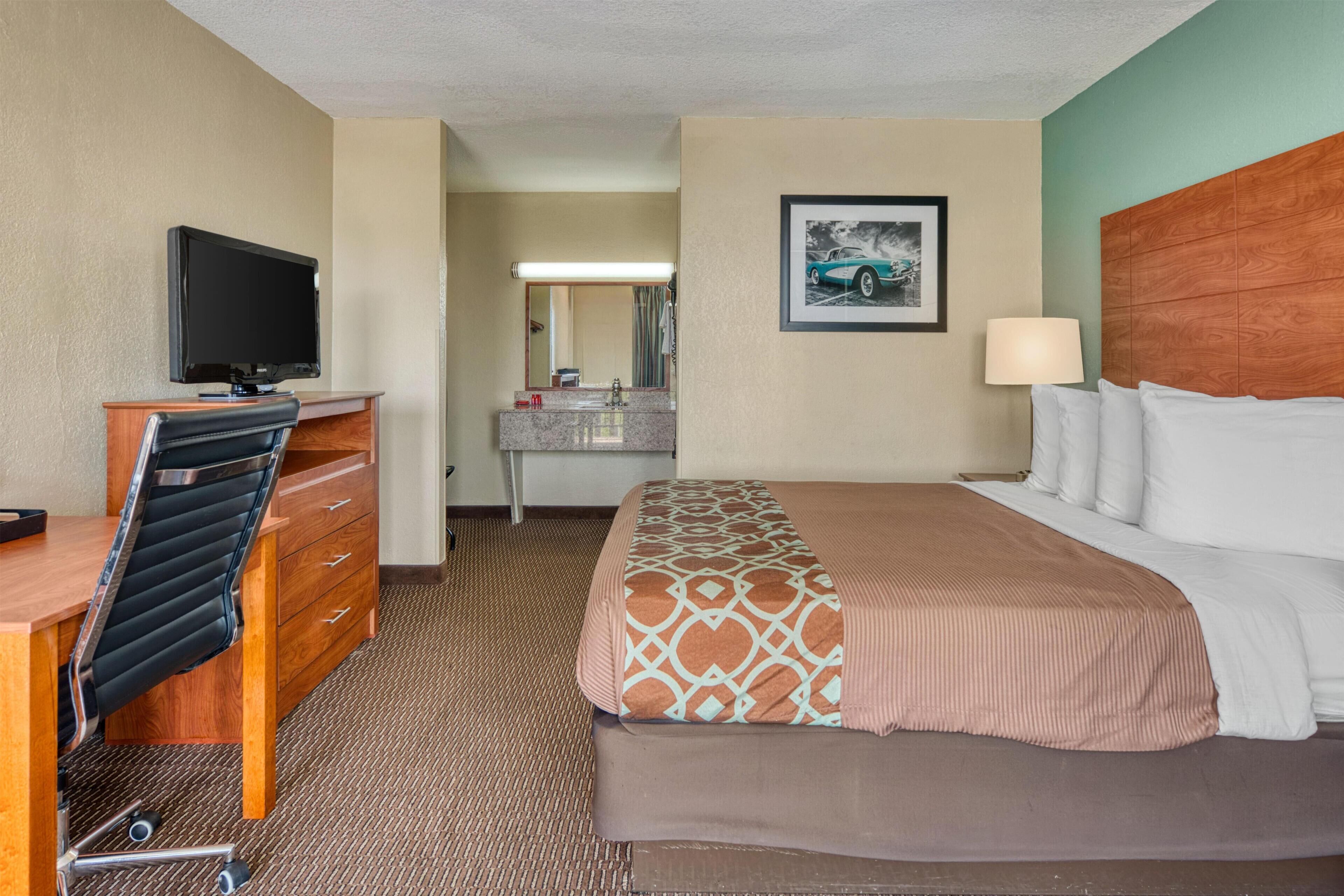 A hotel room with a bed, a desk with a chair, a television, a bathroom with a sink and mirror, and a framed picture on the wall.
