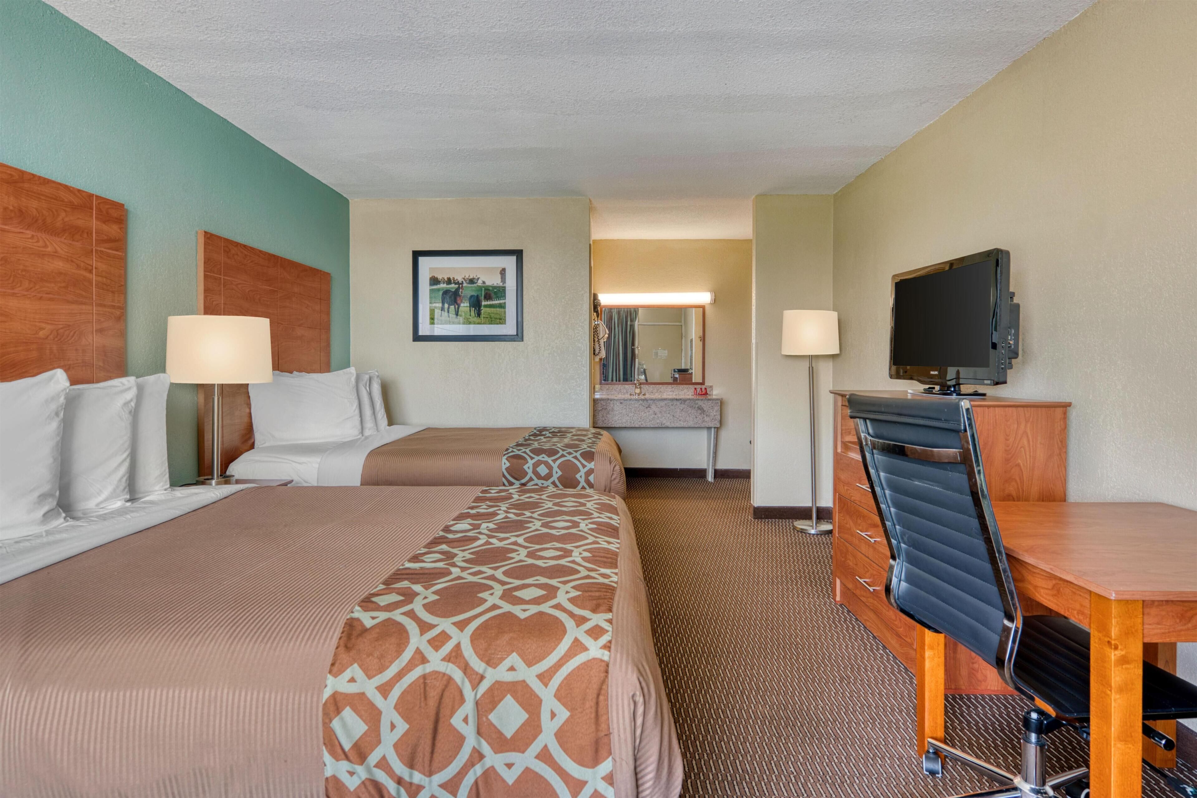 A hotel room with two beds, a desk, a chair, a television, and a bathroom visible in the background.