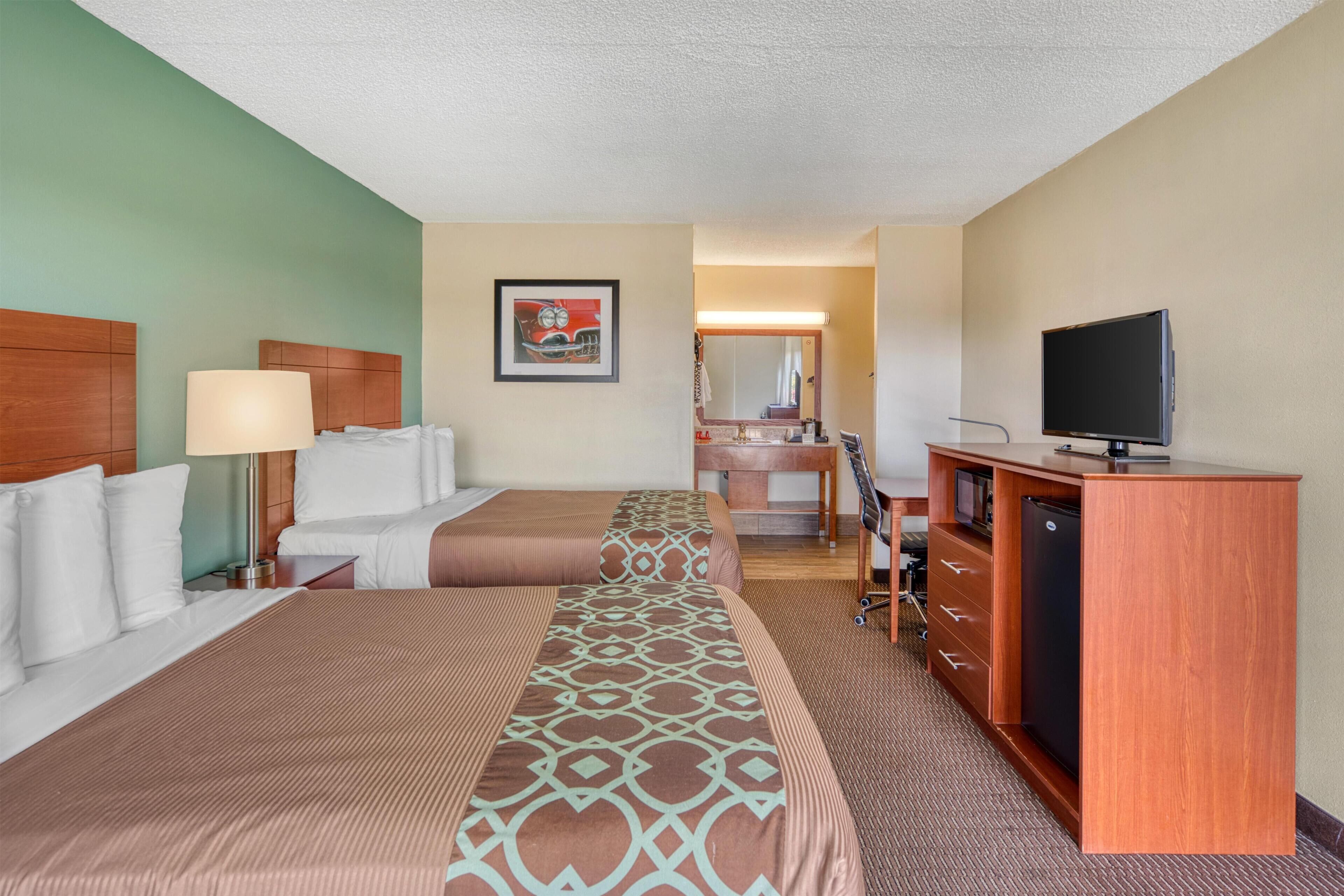 A hotel room with two beds, a desk, a television, a mirror, and a bathroom visible in the background.