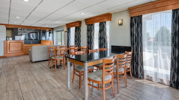 A dining area with a rectangular table, six wooden chairs, a sofa, a kitchen area, and large windows with patterned curtains. - SureStay Hotel by Best Western Bowling Green North (Bowling Green)