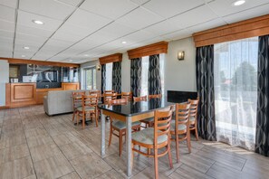 A dining area with a rectangular table, six wooden chairs, a sofa, a kitchen area, and large windows with patterned curtains.
