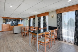 Un comedor con una mesa rectangular, seis sillas de madera, un sofá, una zona de cocina y ventanas grandes con cortinas estampadas.