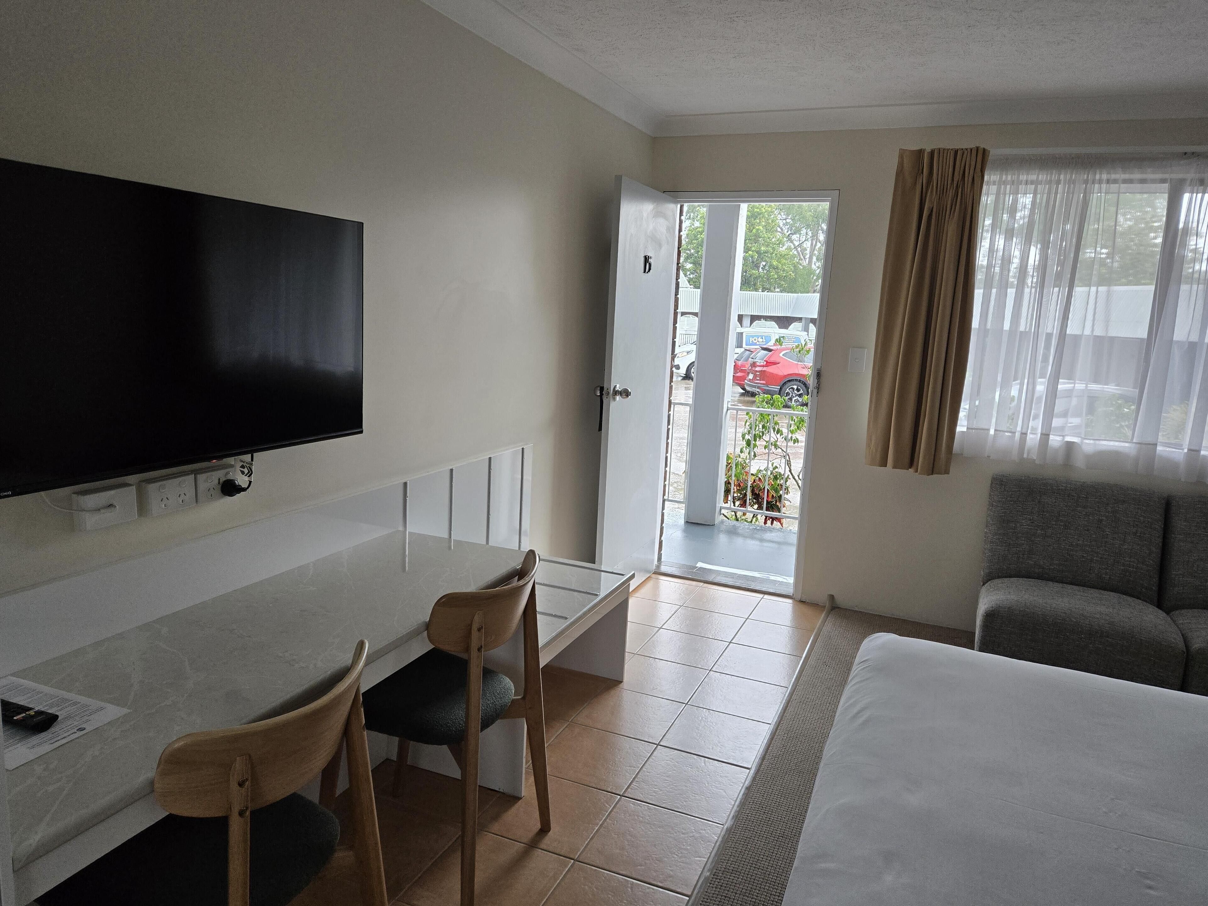 A hotel room with a bed, a desk with a chair, a TV mounted on the wall, and a view of the outside through a door.