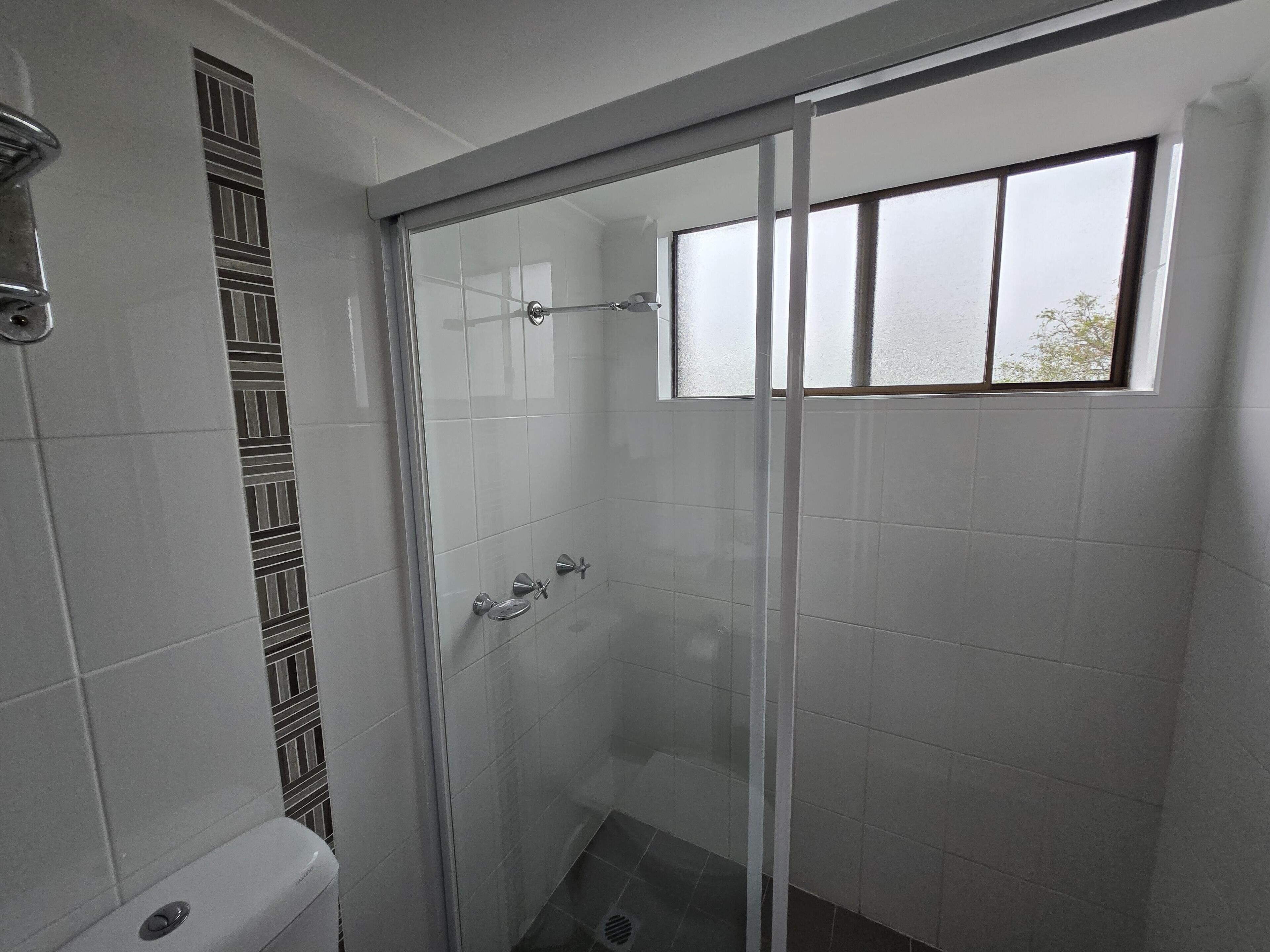 A glass-enclosed shower with a window, white tiled walls, and a toilet with a patterned tile design.