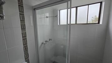 A glass-enclosed shower with a window, white tiled walls, and a toilet with a patterned tile design.