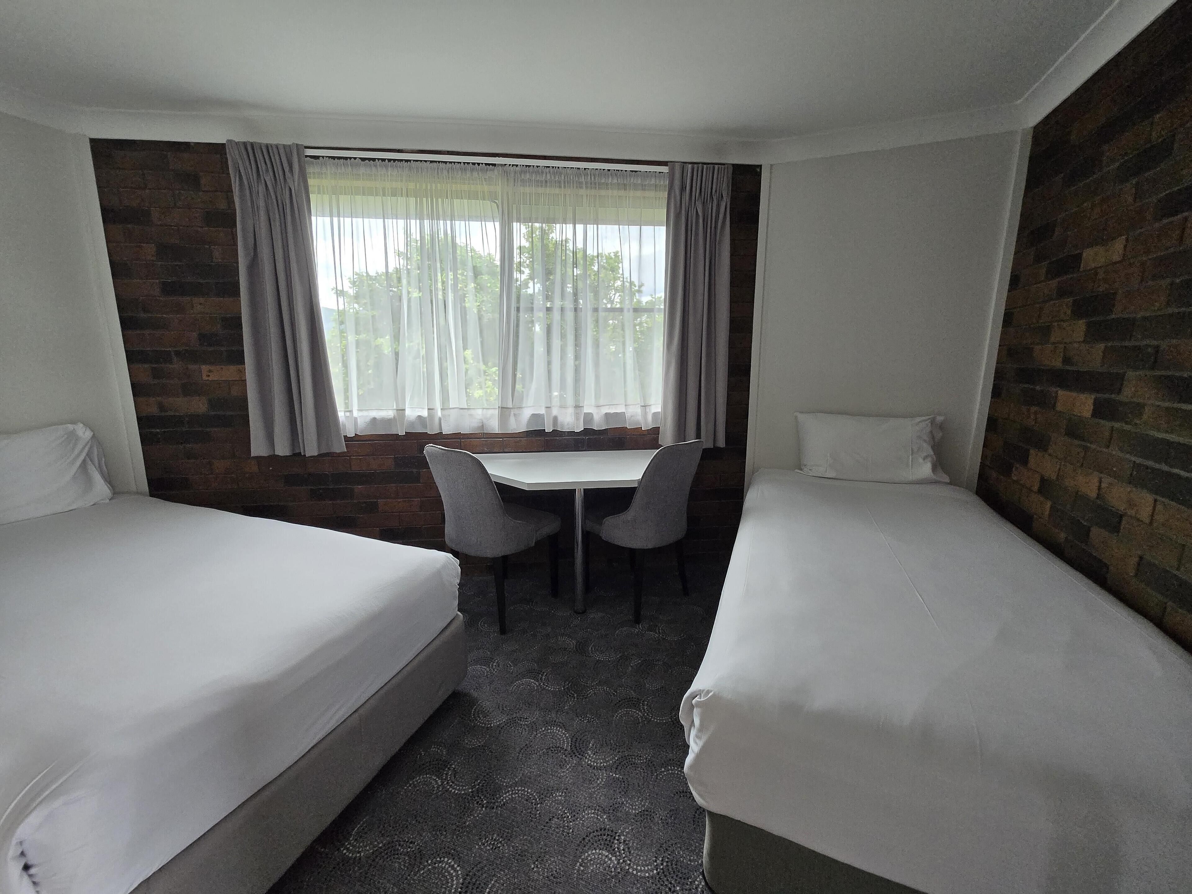 A hotel room with two single beds, a small table with two chairs, a window with curtains, and a brick wall.