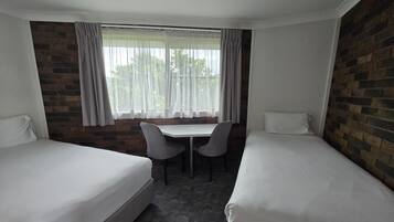 A hotel room with two single beds, a small table with two chairs, a window with curtains, and a brick wall.