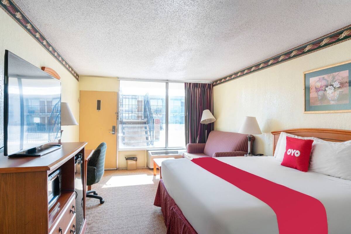 A hotel room with a bed, a desk, a chair, a television, a window with a view of a building, and a painting on the wall.