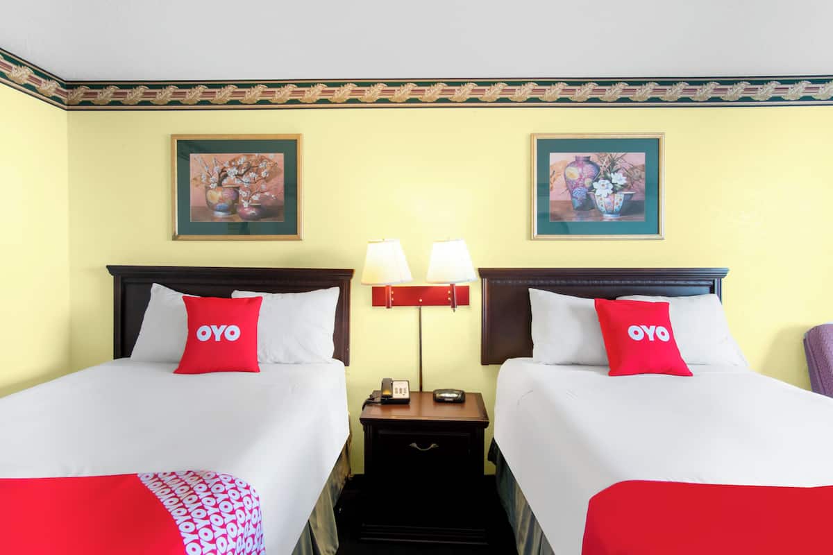 A hotel room with two beds, each with a red pillow labeled "OYO," a bedside table with a lamp, and framed artwork on the walls.