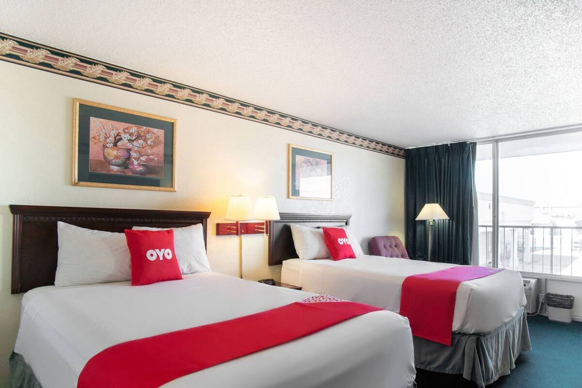 A hotel room with two beds, each with a red OYO pillow, a framed picture above the beds, and a window with curtains.