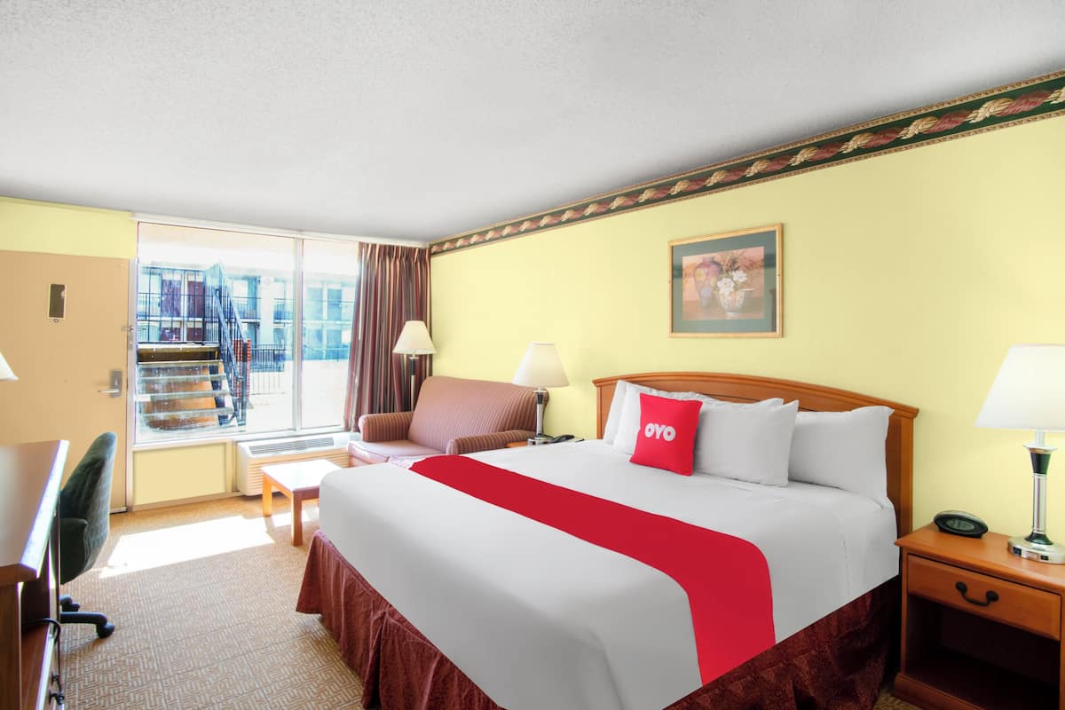 A hotel room with a large bed, a desk, a chair, a nightstand, and a window with a view of a building.
