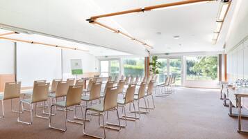 A conference room with rows of chairs, a long table with chairs, and large windows offering a view of greenery outside.