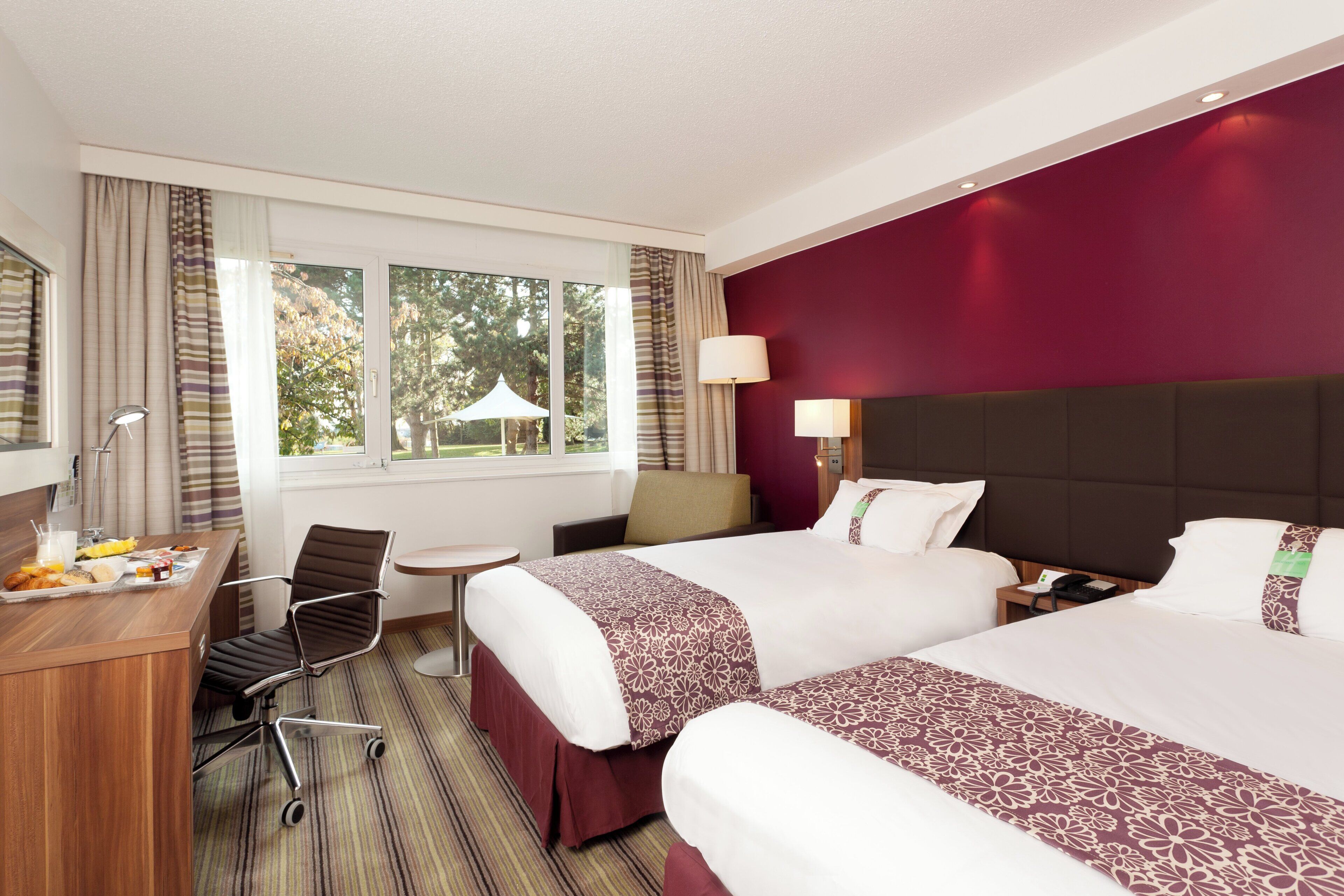 A hotel room with two beds, a desk with breakfast, a chair, and a view of trees through the window.