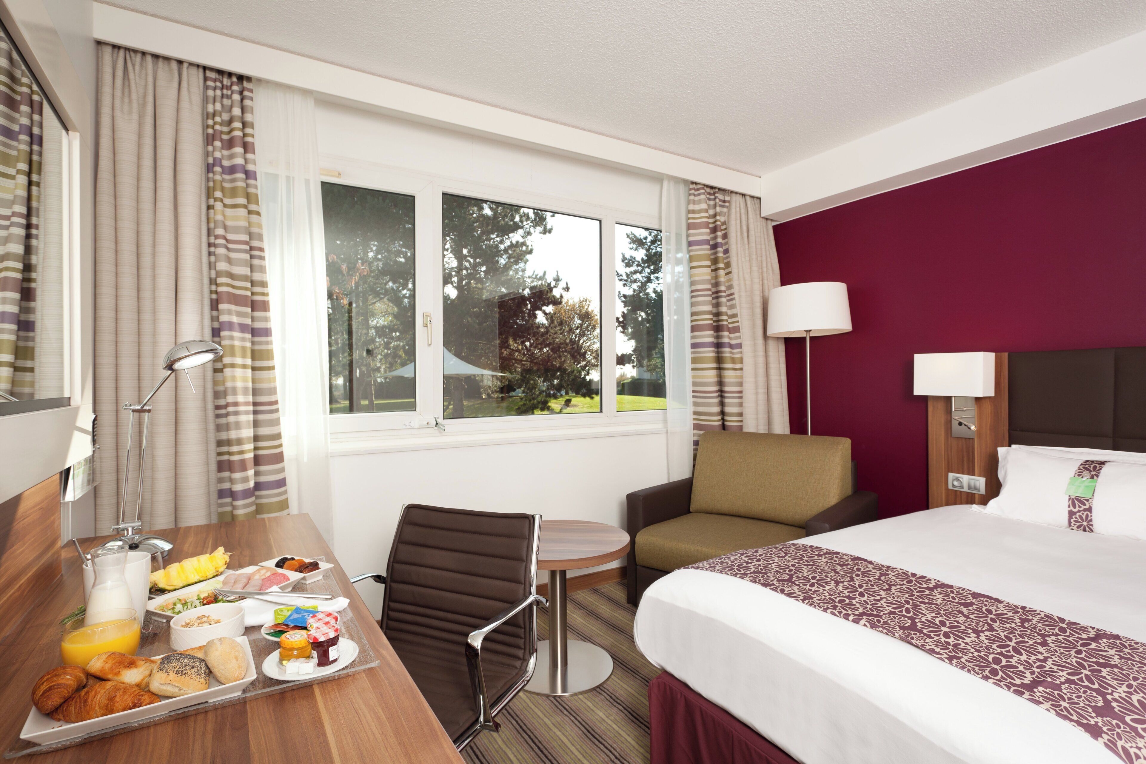 Habitación de hotel con cama, escritorio con desayuno, una silla, lámpara y ventana con vista a árboles.