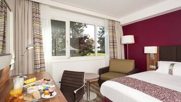Habitación de hotel con cama, escritorio con desayuno, una silla, lámpara y ventana con vista a árboles.