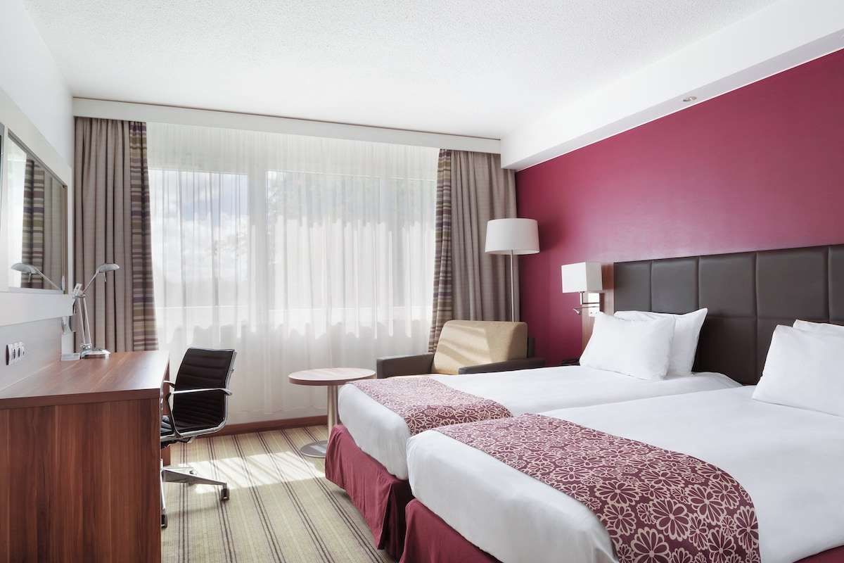 Une chambre d’hôtel avec deux lits, un bureau, une chaise, une lampe et une fenêtre avec des rideaux.