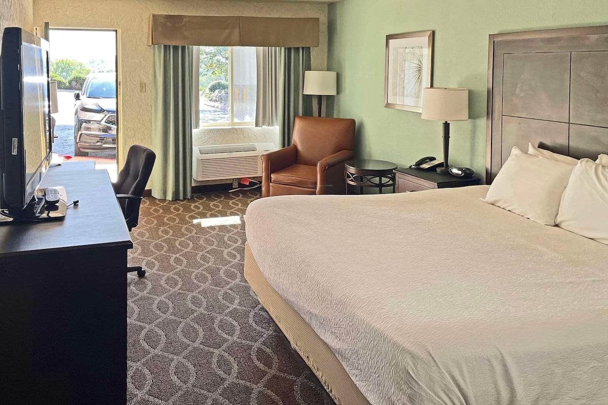 A hotel room with a large bed, a desk with a telephone, a chair, a small table, a lamp, and a window with curtains.