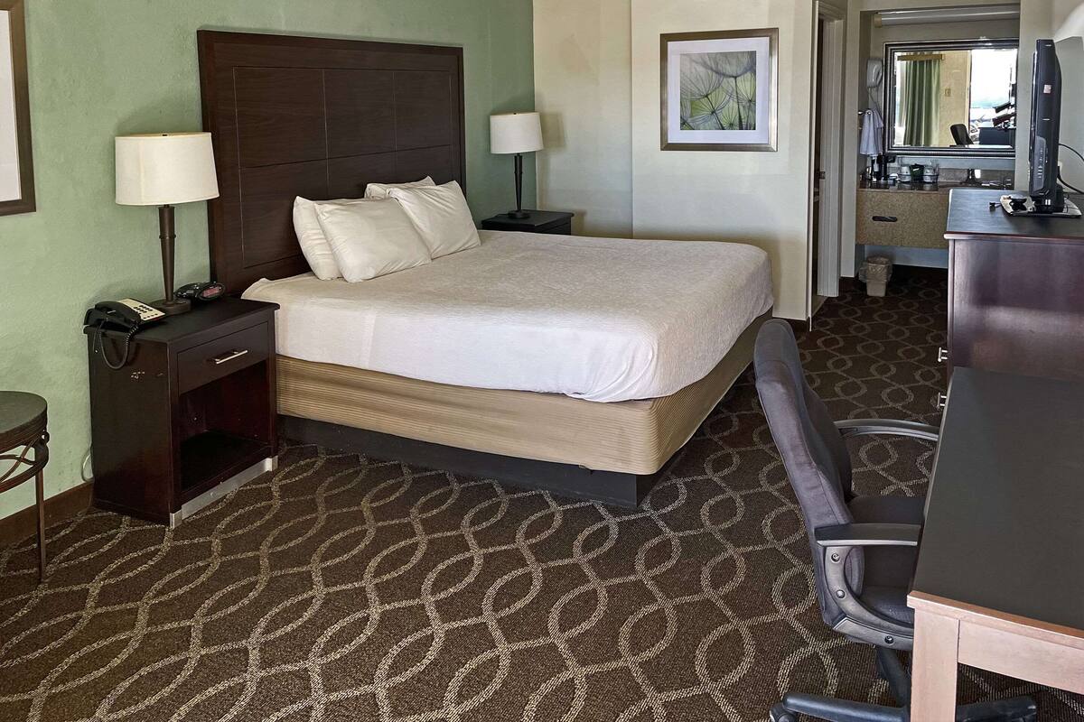 A hotel room with a large bed, bedside lamps, a desk with a computer, and a bathroom visible through an open door.