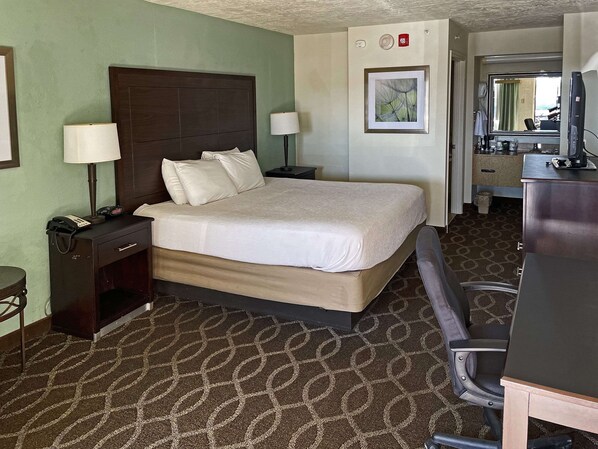 A hotel room with a large bed, bedside lamps, a desk with a computer, and a bathroom visible through an open door.
