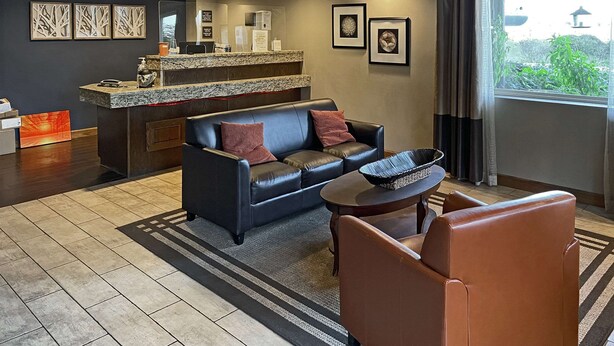 A modern living room with a sofa, armchair, and coffee table. There is a reception desk with a counter and a view of greenery outside.