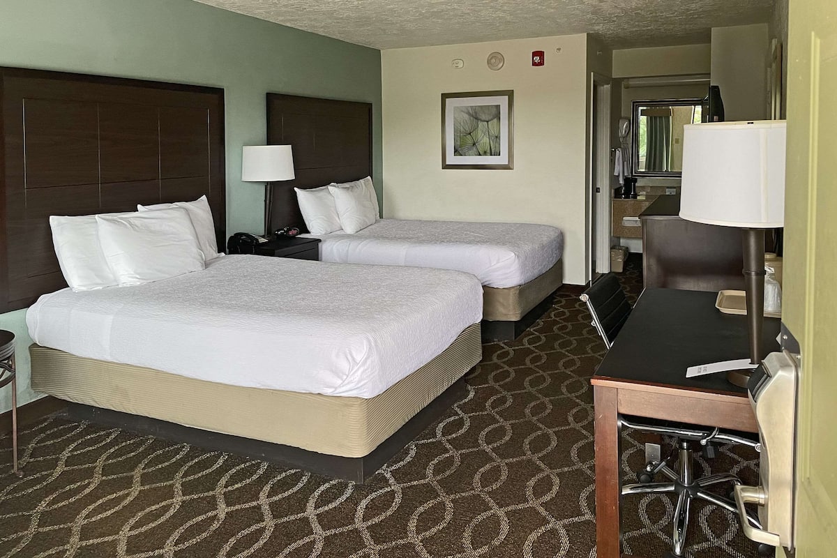 A hotel room with two beds, a desk, a chair, a lamp, a framed picture, and a bathroom visible through an open door.