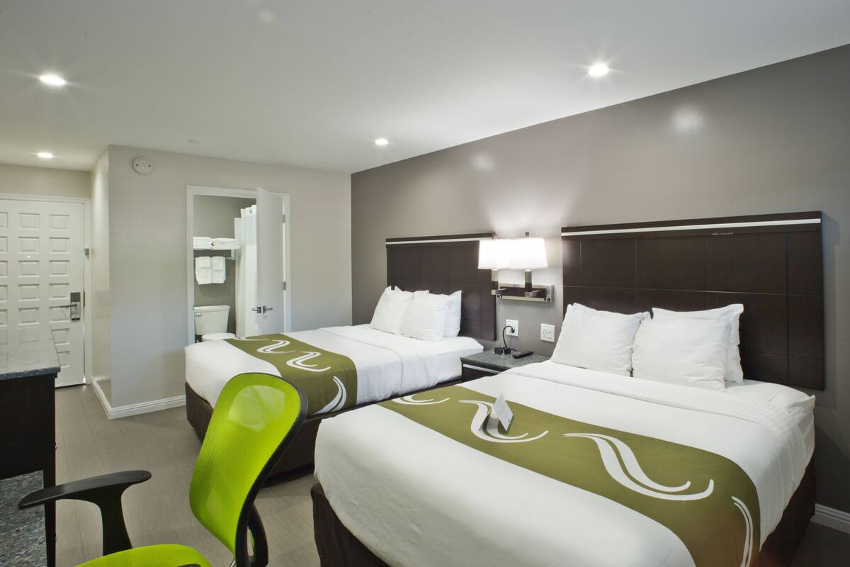 Habitación de hotel con dos camas, un escritorio, una silla y un baño visible a través de una puerta abierta.