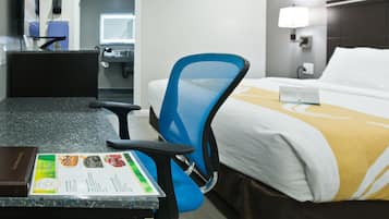 A hotel room with a bed, a desk with a menu, a blue chair, and a bathroom in the background.