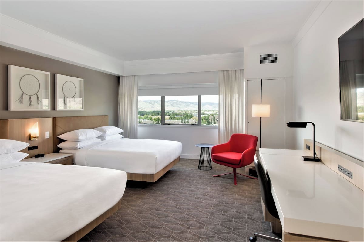A hotel room with two beds, a desk, a red chair, a small table, and a large window with a view of the mountains.