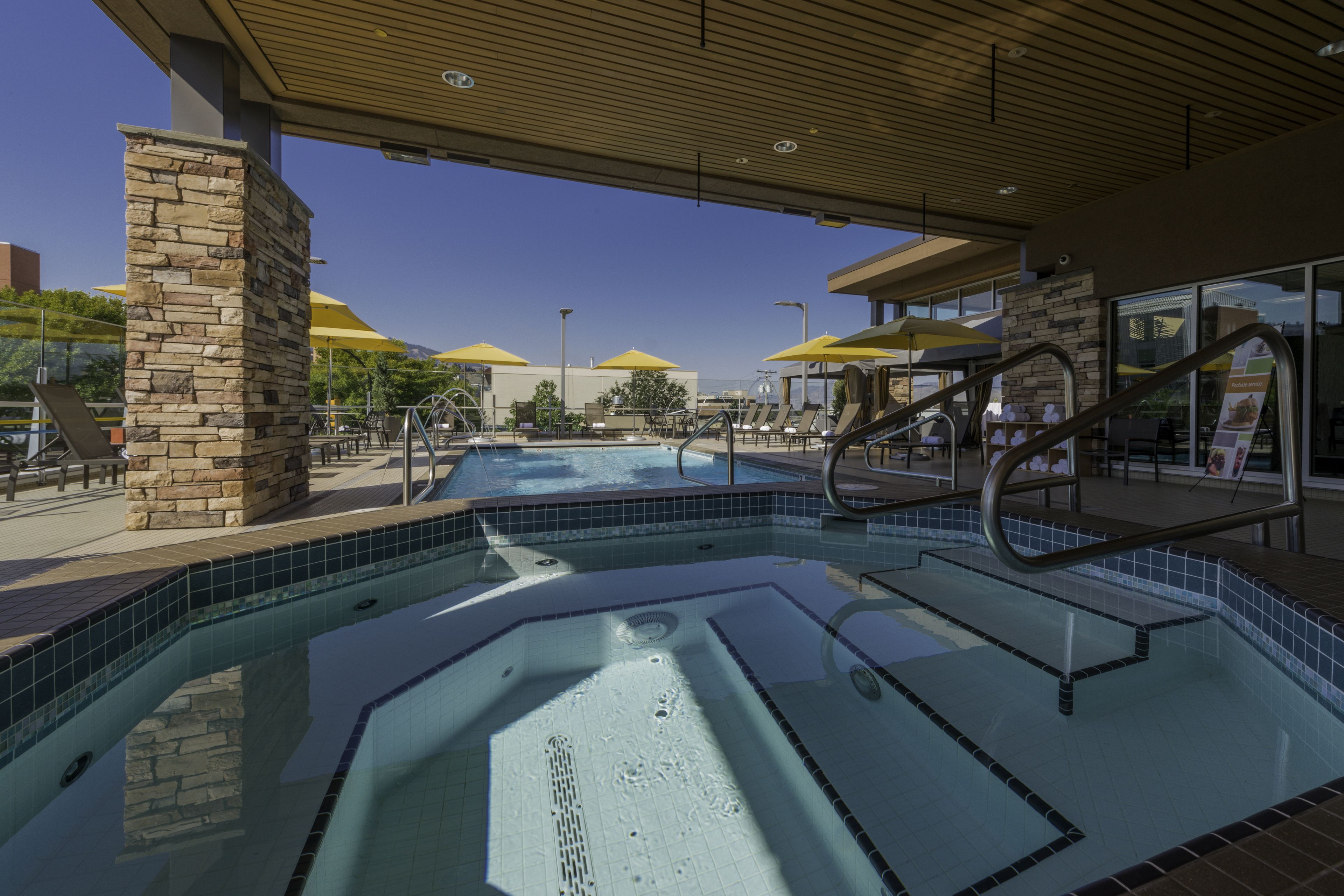 Una piscina con un pilar de piedra, bordes con mosaicos y estructura de techo de madera.