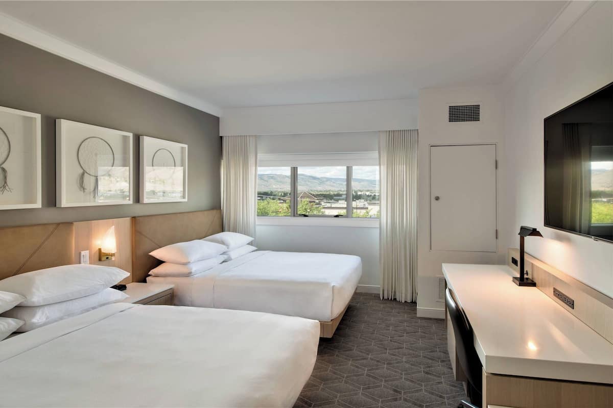 A hotel room with two beds, a desk, a TV, and a window with a view of the mountains.